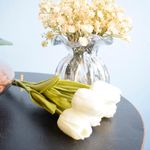 Mini cena. No centro, arranjo artificial de tulipas brancas apoiadas. Ao fundo, vaso redondo de vidro transparente preenchido com flores campestres na cor creme. Ambos apoiados em mesa redonda preta com fundo azul