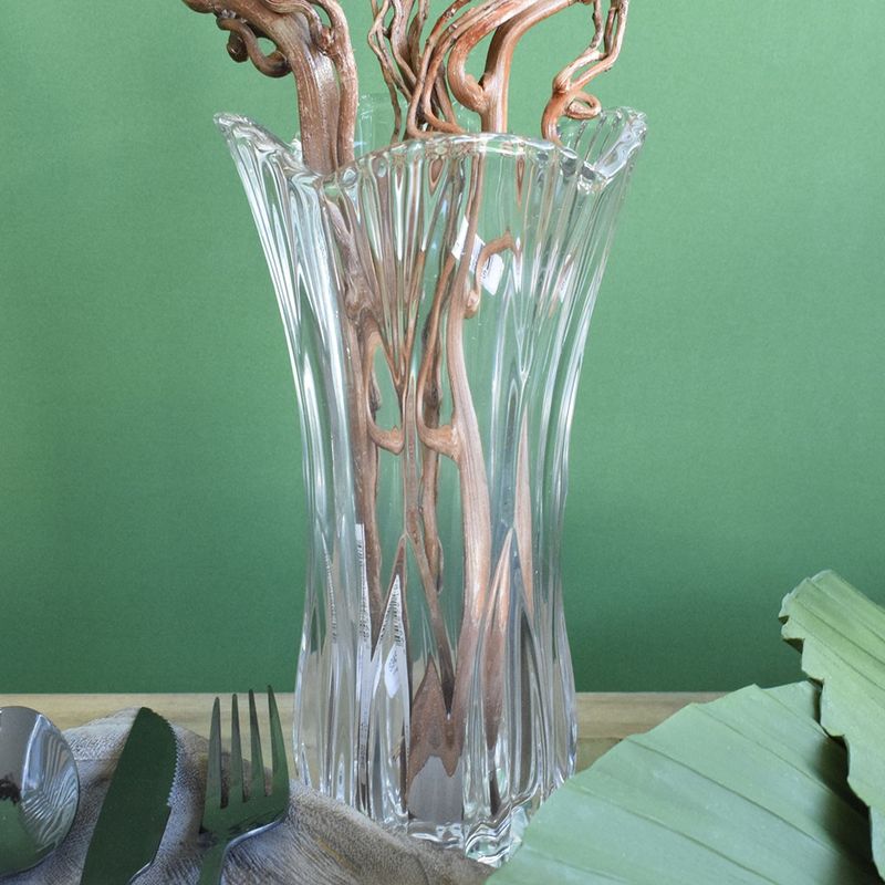 Mini cenário. No centro, vaso de vidro transparente em formato de tulipa preenchido com hastes marrom, no lado esquerdo talheres, no lado direito folhas de latania verde. Todos os itens em fundo verde