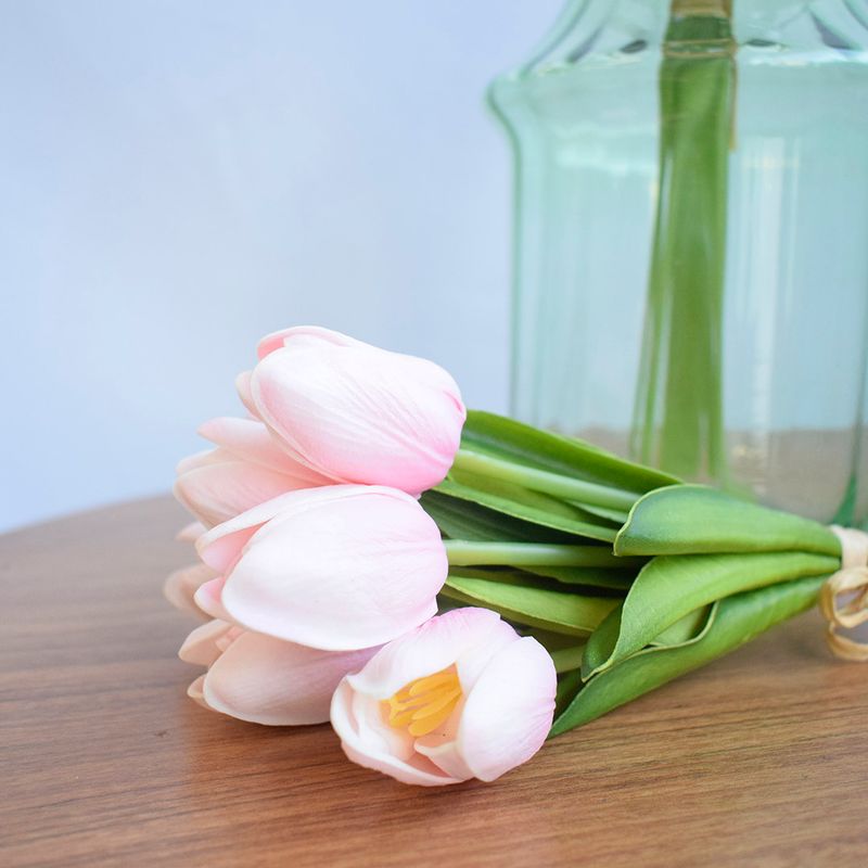 No lado direito, vaso de vidro transparente preenchido com hastes verdes. Na frente, buque de mini tulipas rosa claro. Ambos os itens em mesa de madeira redonda em fundo azul