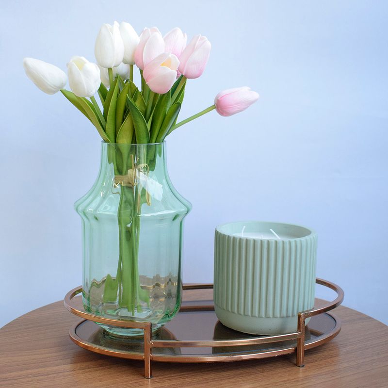 Mini cenário. No centro. arranjo de tulipas rosas e brancas em vaso de vidro transparente. Lado direito, vela em  cerâmica azulada. Ambos os itens sobre bandeja metalica vazada apoiada em mesa redonda de madeira em fundo azul