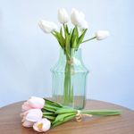 No centro, arranjo de tulipa branca em vaso de vidro transparente, frente ao vaso buque de tulipas rosa claro. Ambos os itens sobre mesa redonda de madeira em fundo azul