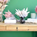 Mini cena. Aparador de madeira com decoração em folhagens artificiais rosa, pink e creme apoiado sobre caixa livro e vaso trouxinha murano preto em fundo verde