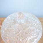 No centro, foco aproximado da tampa da bomboniere de vidro com acabamento em desenho de cristais e estrelas sobre mesa de madeira em fundo azul