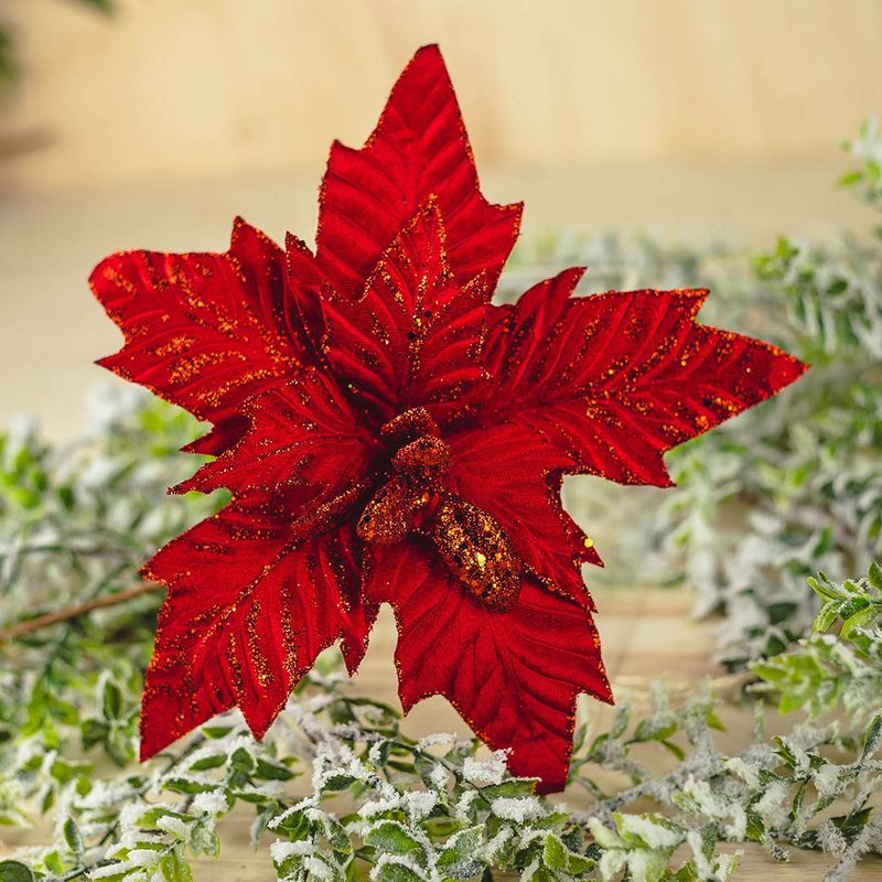 No centro, flor de natal em veludo vermelho com detalhes em glitter vermelho no fundo de madeira decorado com folhagens nevada