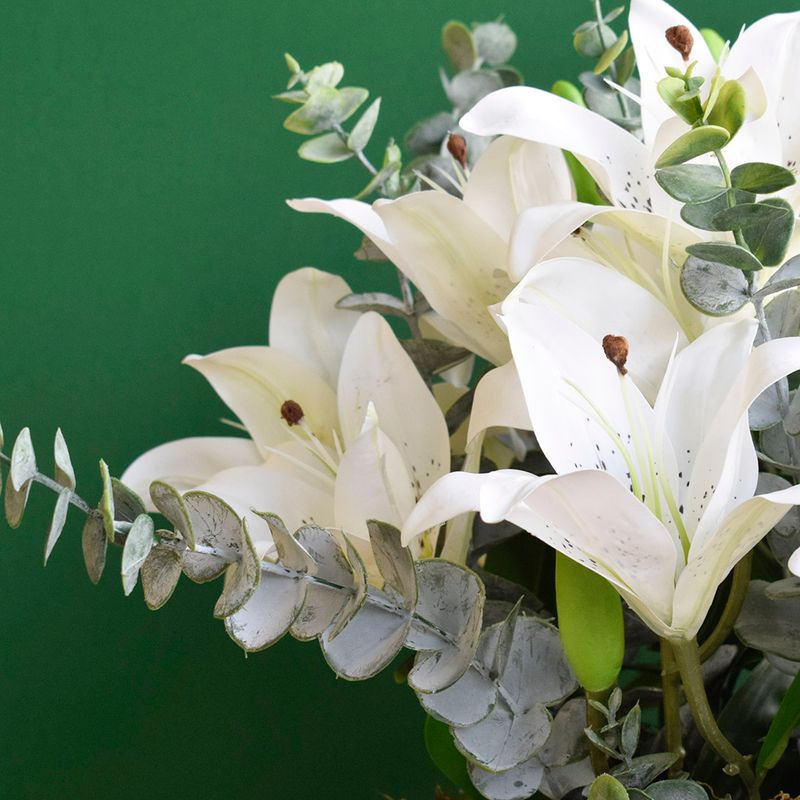 Na lateral direita, flores do arranjo artificial de lirio branco com ramos de eucalipto em fundo verde