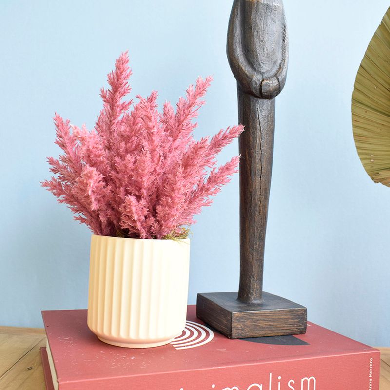 Mini cenário. Lado esquerdo, arranjo de flores secas pink em vaso canelado areia. Lado direito, estátua humana de madeira. Itens sobre caixa livro em fundo azul