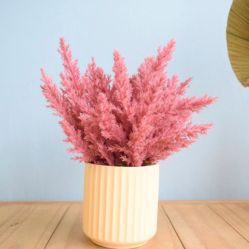 No centro, arranjo de flores secas na cor pink em vaso canelado areia sobre mesa de madeira em fundo azul