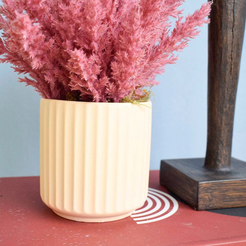 Lado esquerdo, foco aproximado da lateral do vaso canelado areia com arranjo de flores secas pink. Lado direito, adorno tipo estatua marrom. Itens apoiados sobre caixa livro em fundo azul