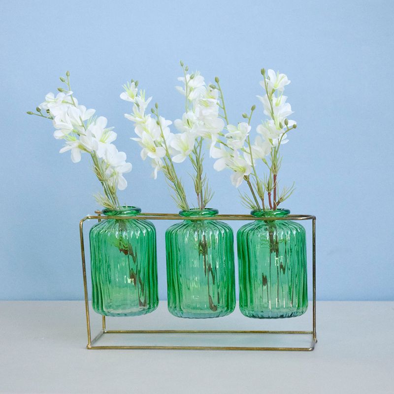 No centro, trio de vasos tipo garrafinhas verdes em suporte metalico preenchidas com arranjo de flor campestre em fundo azul