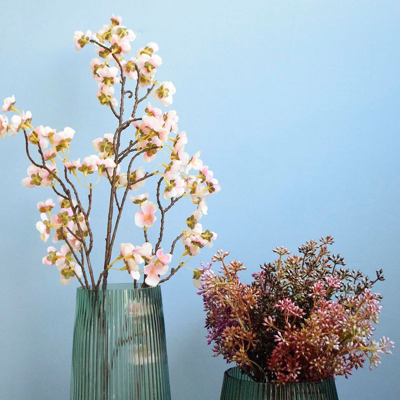 Lado esquerdo, haste de flor de cerejeira rosa claro em vaso de vidro canelado. Lado direito, buque de sementinha em vaso de vidro canelado. Itens em fundo azul