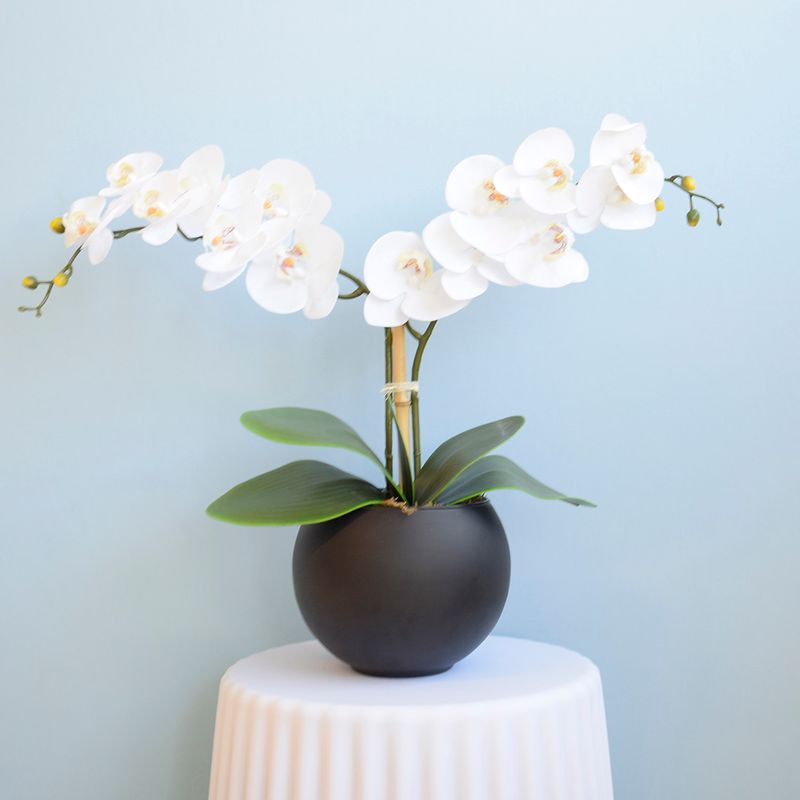 No centro, arranjo artificial com duas hastes de orquidea branca no vaso redondo preto fosco apoiado sobre mesa lateral coberta com toalha branca em fundo azul