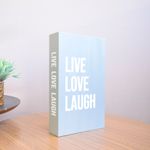 No centro, na vertical caixa livro cinza com tipografia branca escrita "Live Love Laugh" apoiado sobre mesa redonda de madeira em fundo rose