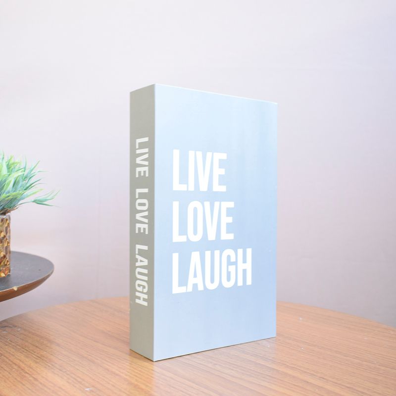 No centro, na vertical caixa livro cinza com tipografia branca escrita "Live Love Laugh" apoiado sobre mesa redonda de madeira em fundo rose