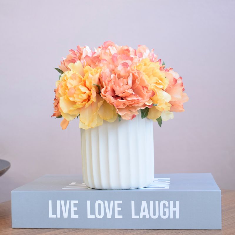 Mini cena. No centro, arranjo de rosas amarelas e rosas em vaso de ceramica canelada branca apoiada sobre caixa livro cinza com tipografia branca escrita "live, love laugh" em fundo rose