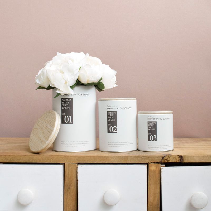 No centro e enfileiradas, 3 latas decorativas brancas com tampa de madeira, uma decoraca com buque de rosas, todas apoiadas sobre aparador de madeira com gavetas brancas em fundo rose