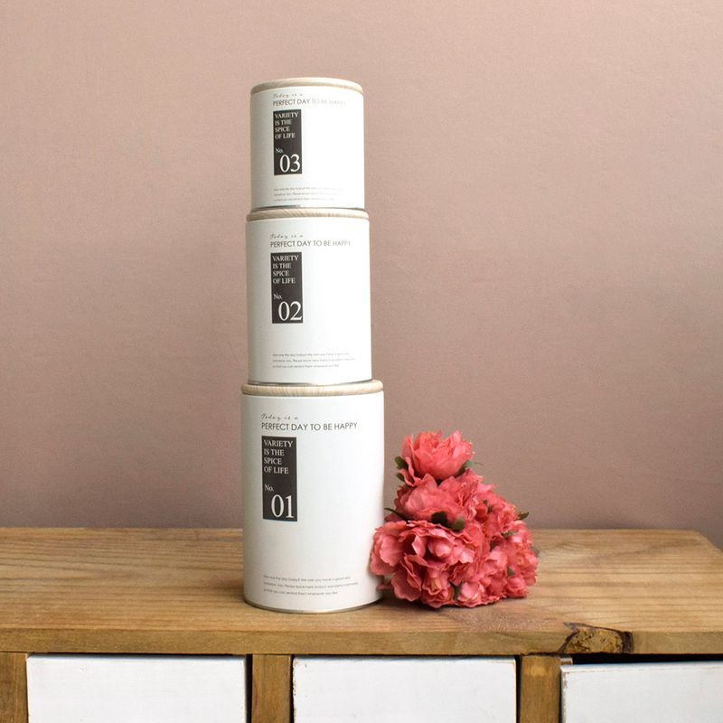 No centro e empilhadas, 3 latas decorativas brancas com tampa de madeira e tamanhos diferentes, apoiadas sobre aparador de madeira com gavetas brancas em fundo rose.