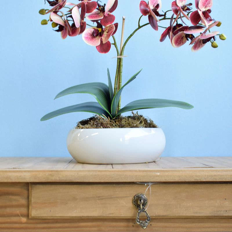 No centro, foco aproximado da lateral do vaso de ceramica branca tipo bacia com arranjo artificial de orquidea marsala. Item apoiado sobre aparador de madeira com gaveta em fundo azul