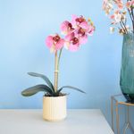 No centro, arranjo de orquidea rosa em vaso canelado creme sobre mesa de madeira branca. No lado direito, arranjo de cerejeira em vaso de vidro transparente apoiado sobre mesa lateral estilo industrial.
