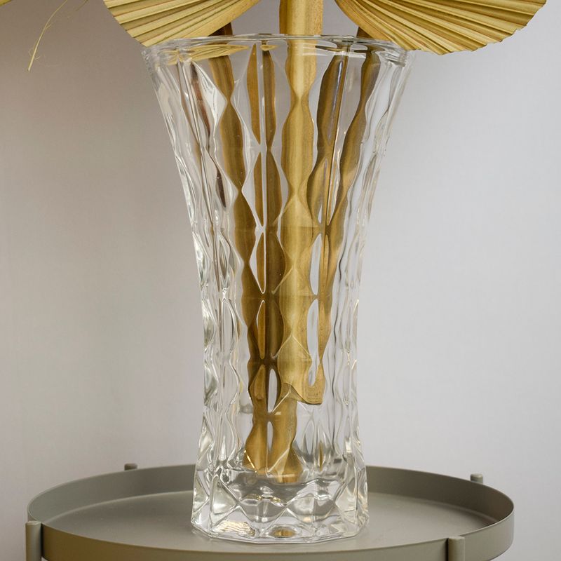 No centro, foco aproximado da lateral do vaso de vidro transparente em formato de tulipa com hastes de latania dentro e apoiado sobre mesa lateral redonda fendi em fundo azul