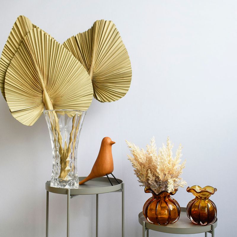 Mini cena. Mesas laterais redondas fendi decoradas com vasos trouxinhas ambar, adorno de passarinho e arranjo de latinia palha em vaso de vidro transparente em formato de tulipa. Itens em fundo azul