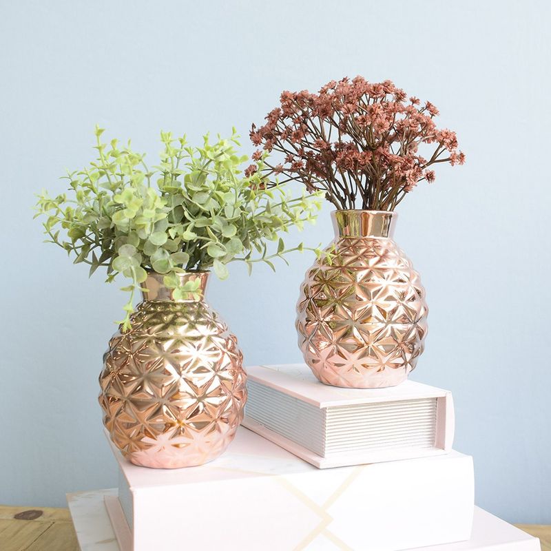 No centro, dois arranjo de eucalipto verde e flor mosquitinho marsala, ambos artificiais em vasos em formato de moringa de vidro espelhado rose gold apoiado sobre caixa livro branca em fundo azul