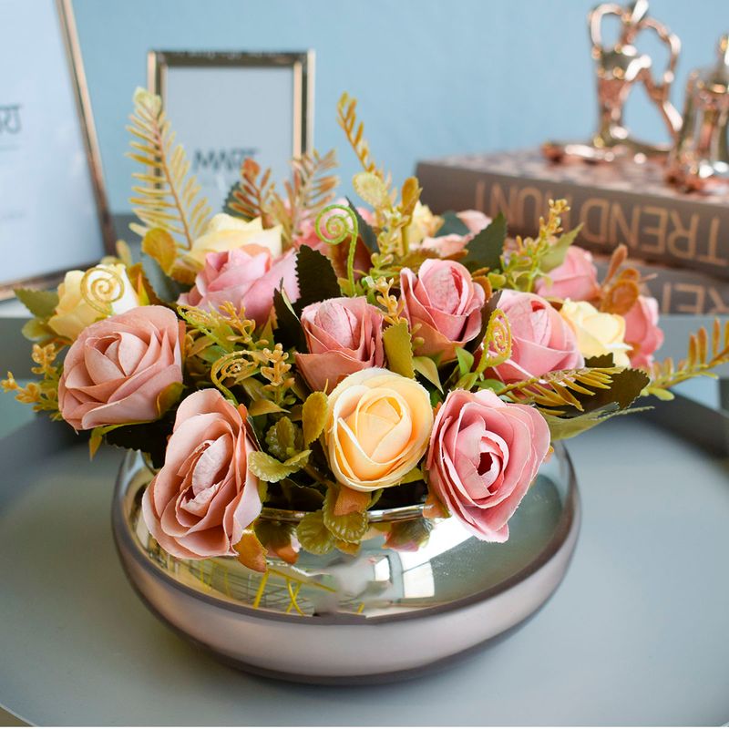 Mini cena. Na frente, arranjo artificial de rosas amarelas e rosas em vaso lagoon rose apoiado em mesa redonda. No fundo, porta retratos metalicos, caixa livro e escultura de yogui. Todos os itens em fundo azul.